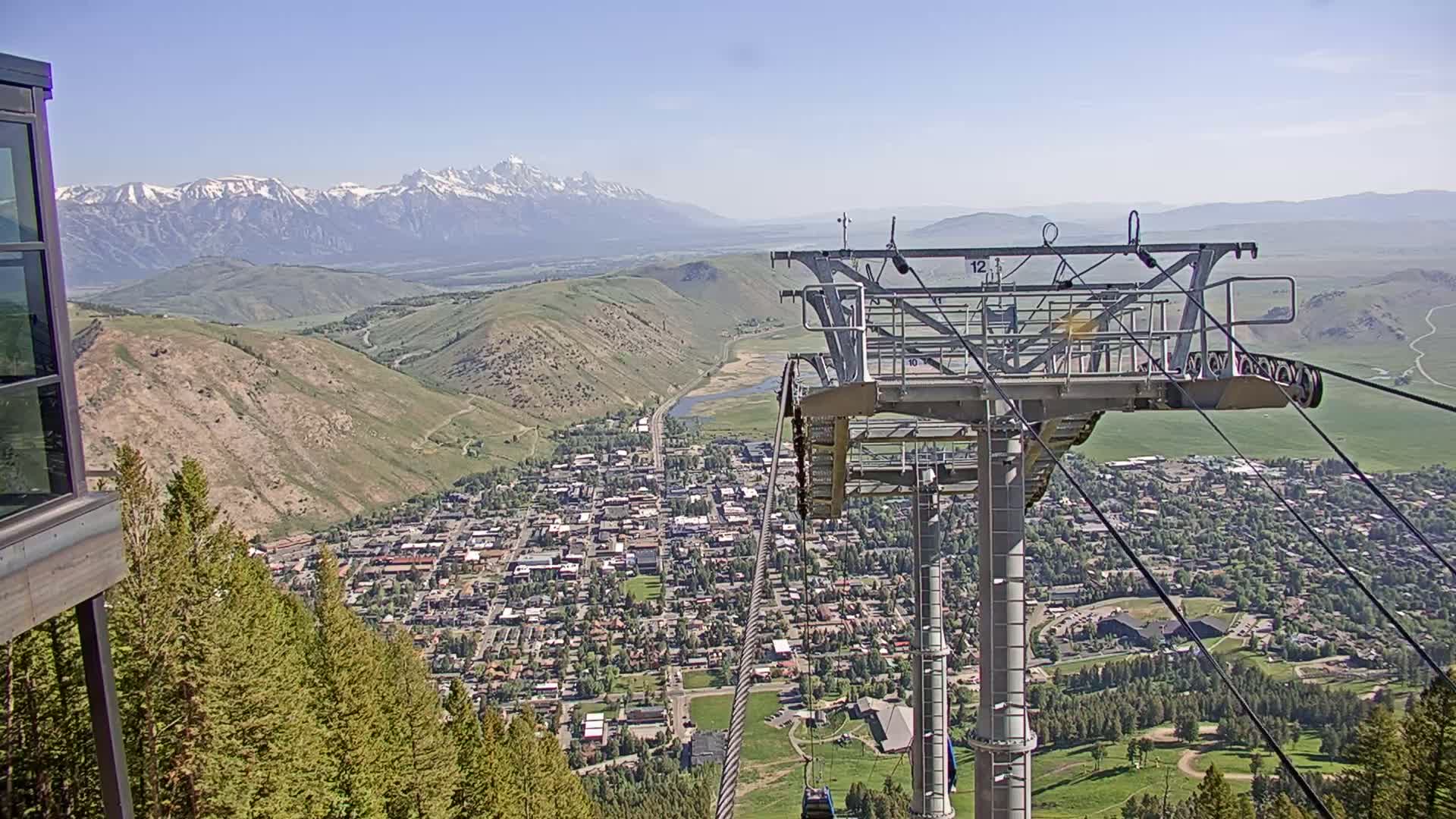 A chairlift ascends a mountain overlooking a valley town and snow-capped mountains under a clear blue sky.
