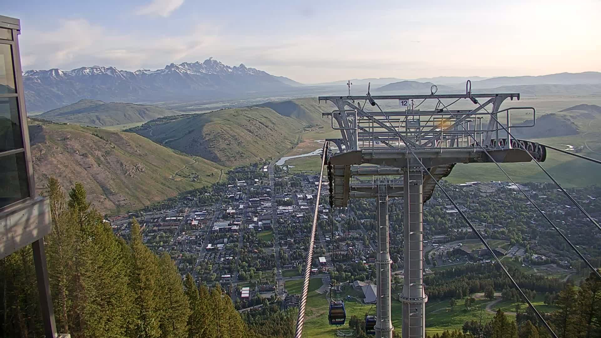 A chairlift ascends a mountain overlooking a valley town and snow-capped mountains under a mostly clear sky.