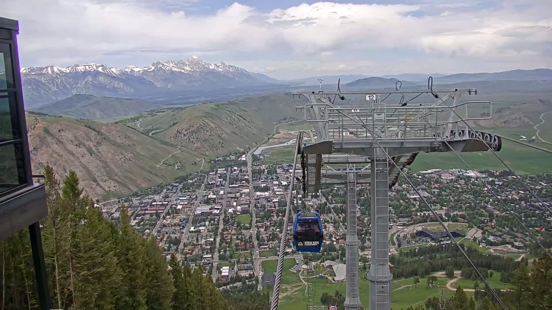 A partly cloudy day shows a mountain town nestled in a valley with snow-capped mountains in the background, viewed from a cable car ascending a mountain.