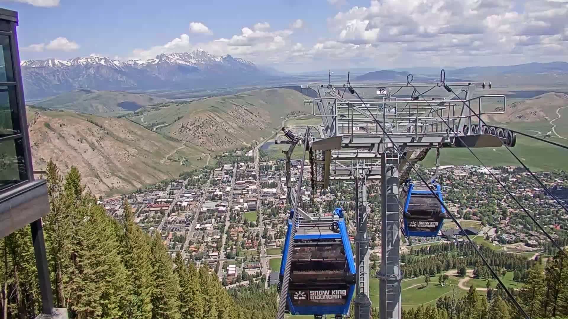 A chairlift ascends a mountain overlooking a town nestled in a valley, with snow-capped mountains in the distance under partly cloudy skies.