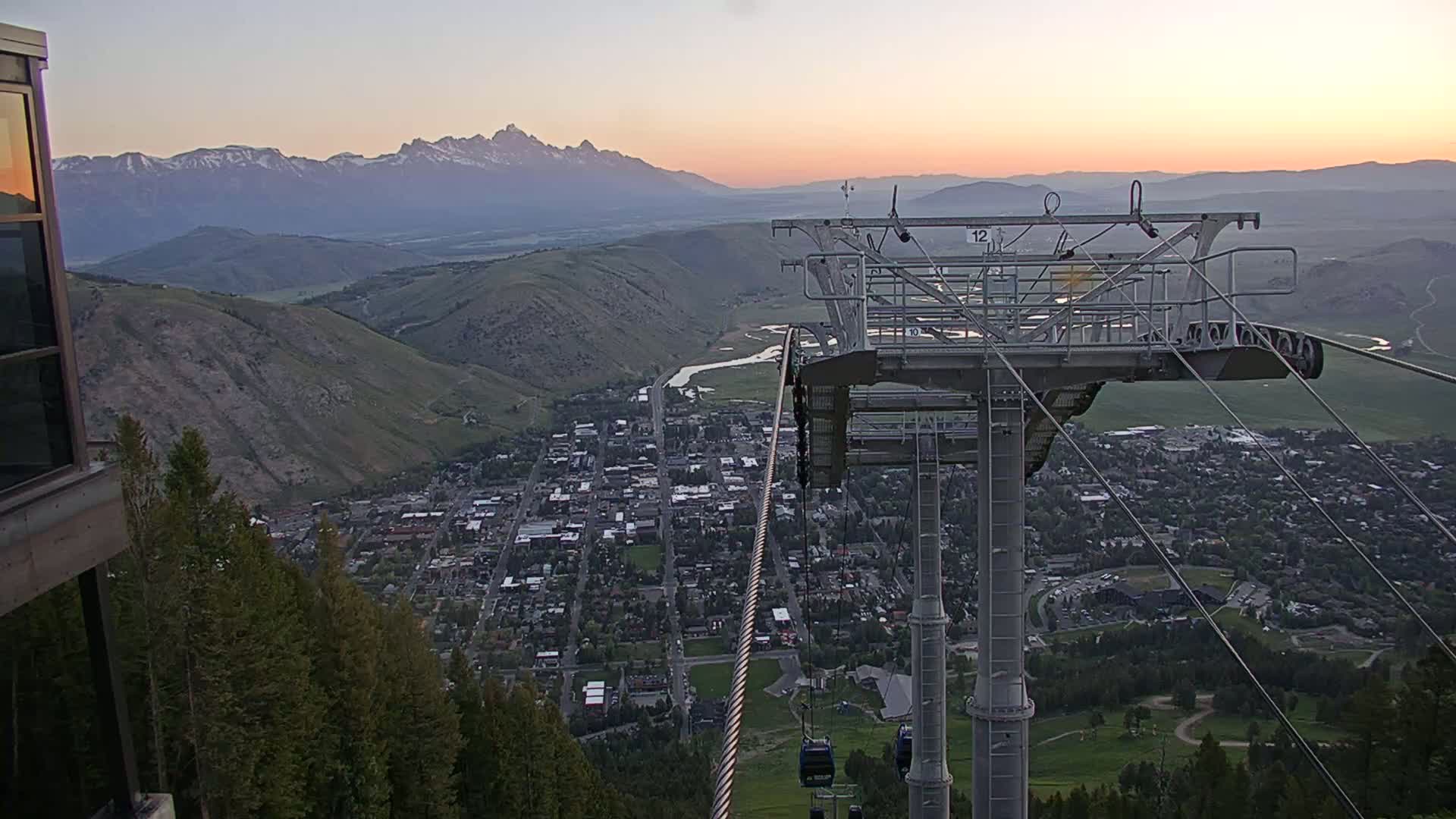 A mountain town at sunset is viewed from a chairlift overlooking a valley and distant snow-capped mountains.
