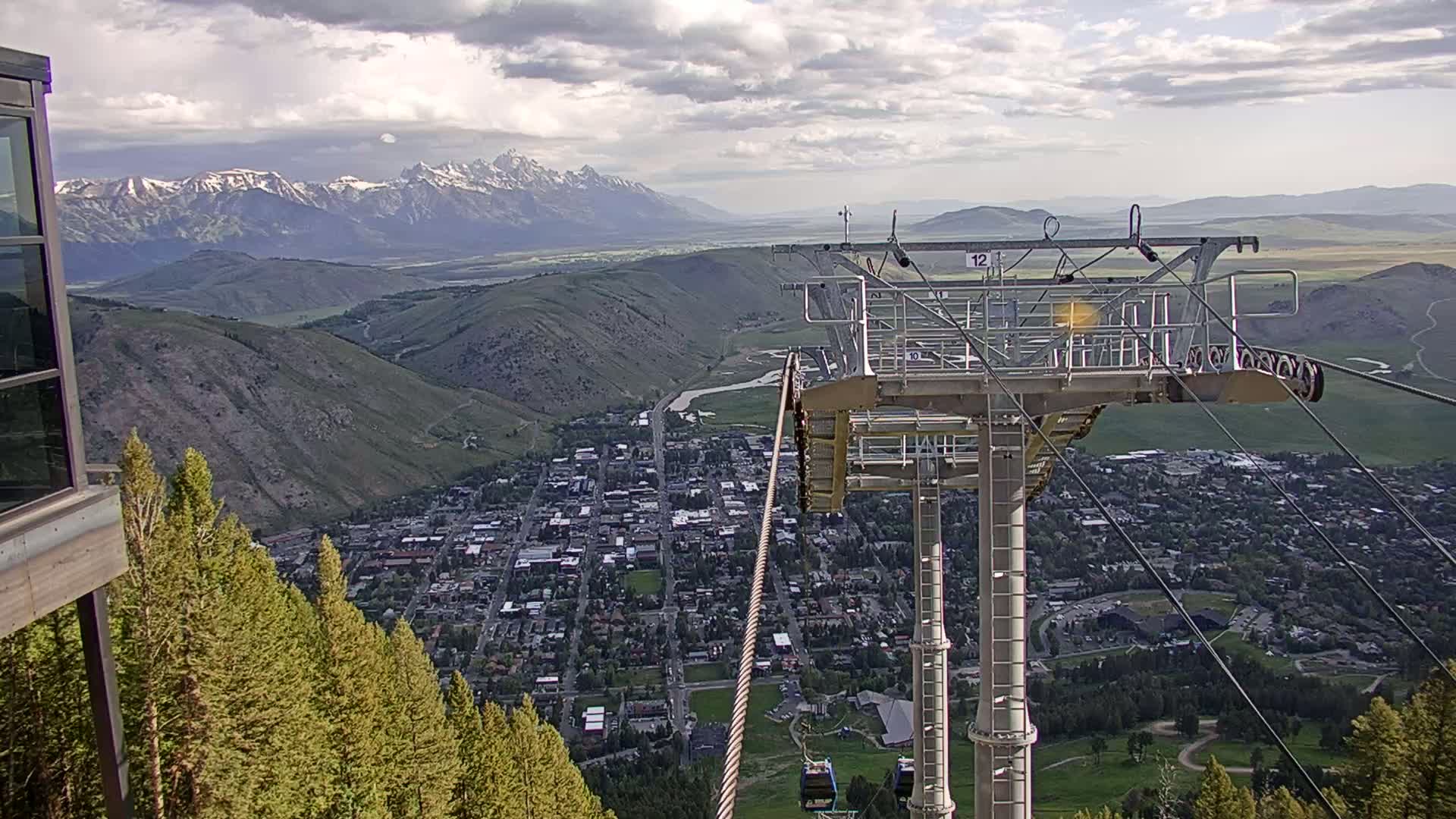 A chairlift ascends a mountain overlooking a town nestled in a valley, with snow-capped mountains in the distance under partly cloudy skies.