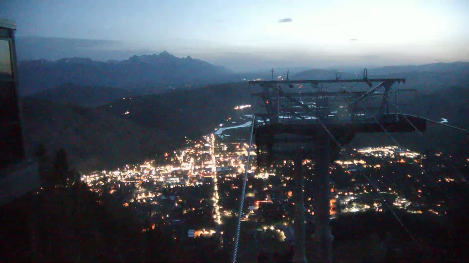 A nighttime view from a chairlift shows a valley town illuminated below, with mountains silhouetted in the distance under a twilight sky.