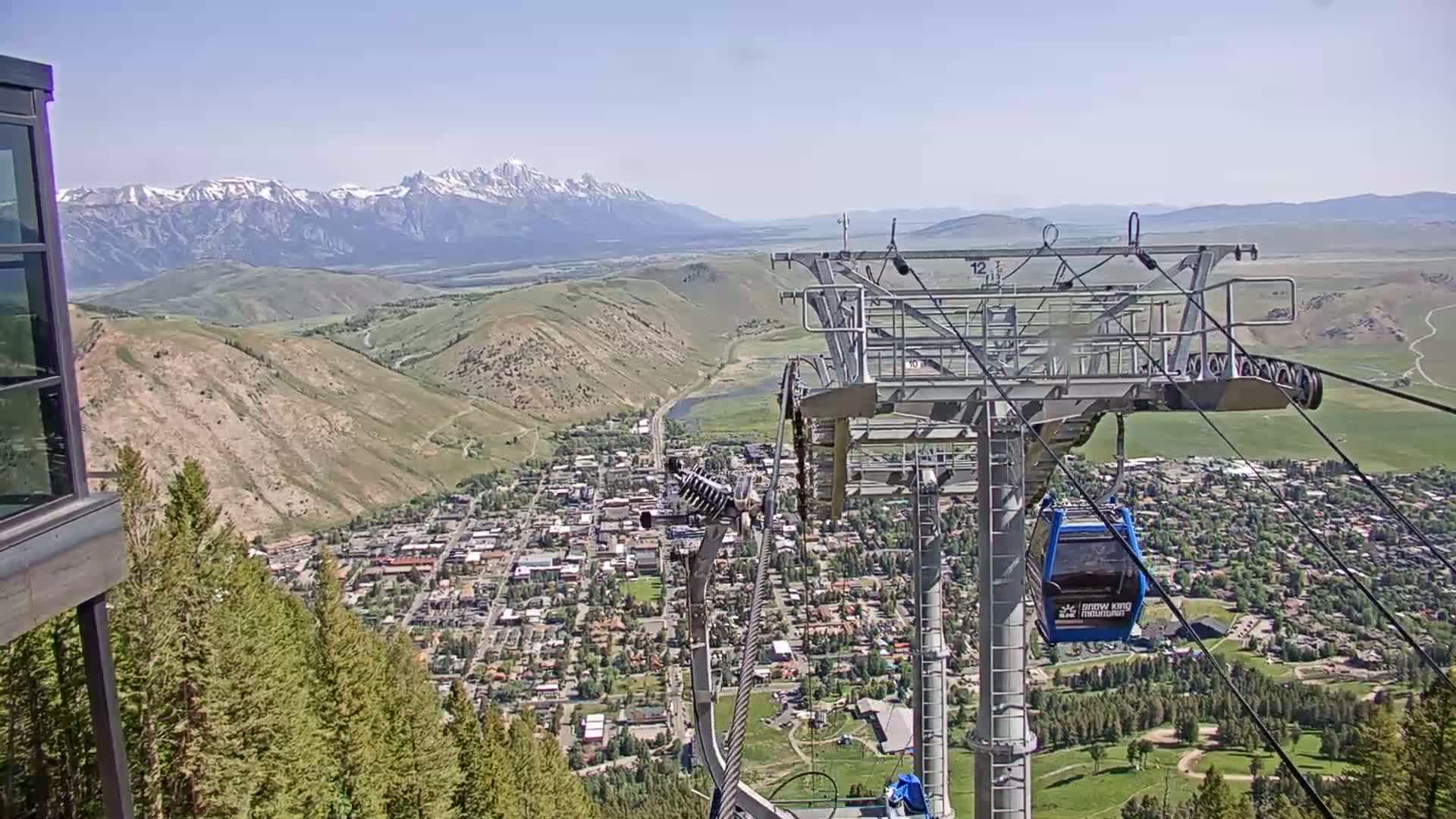 A chairlift ascends a mountain overlooking a town nestled in a valley beneath snow-capped mountains on a clear, sunny day.