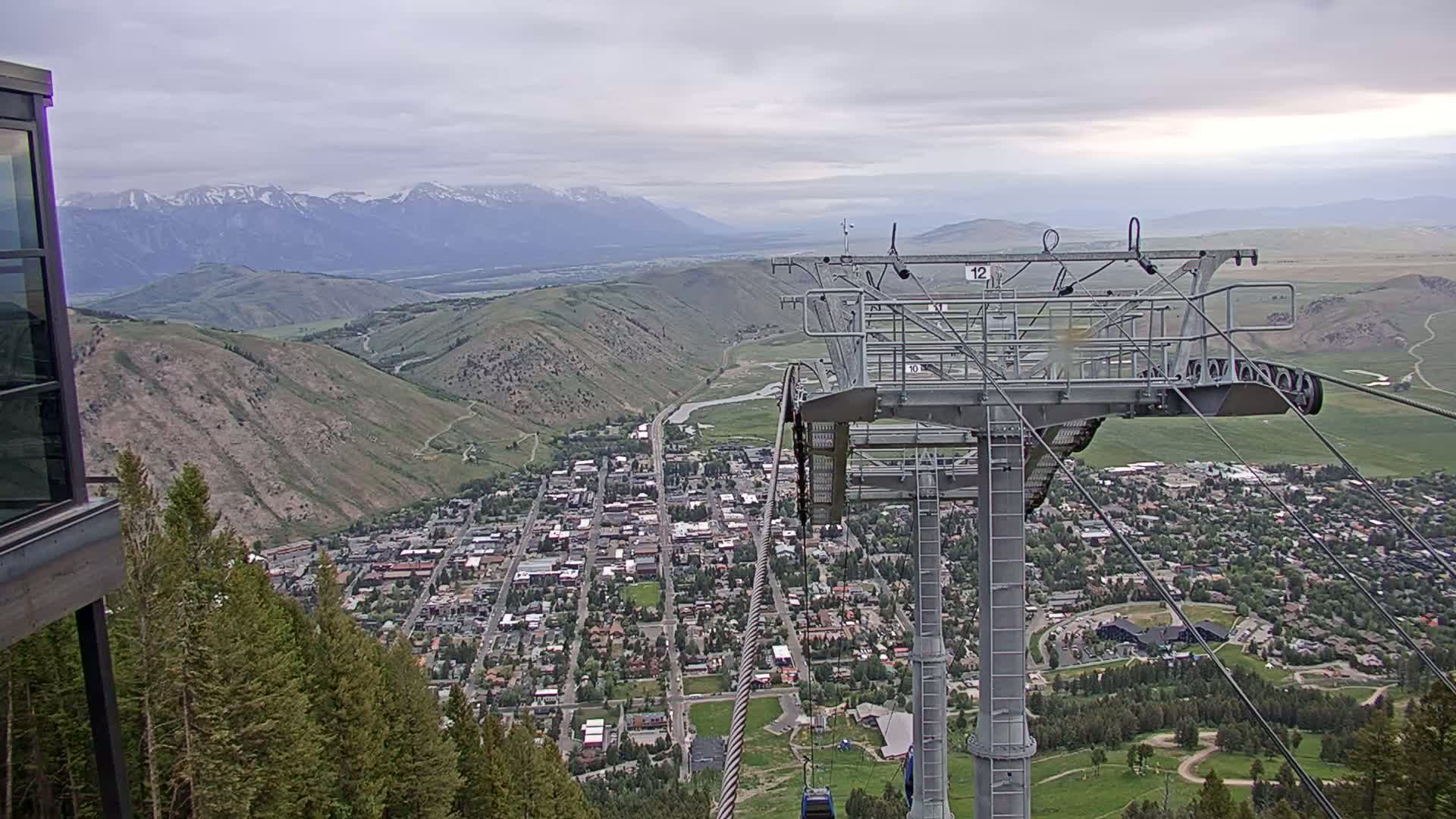 A chairlift overlooks a town nestled in a valley, with snow-capped mountains in the background under an overcast sky.