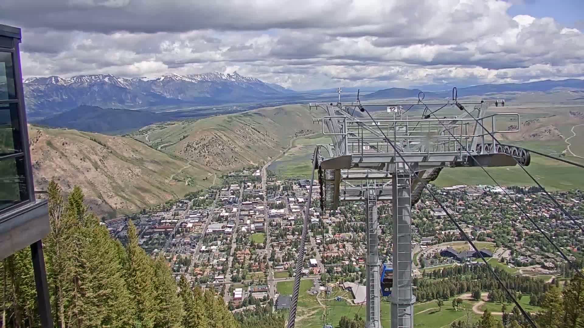 A chairlift overlooks a town nestled in a valley below snow-capped mountains under a partly cloudy sky.