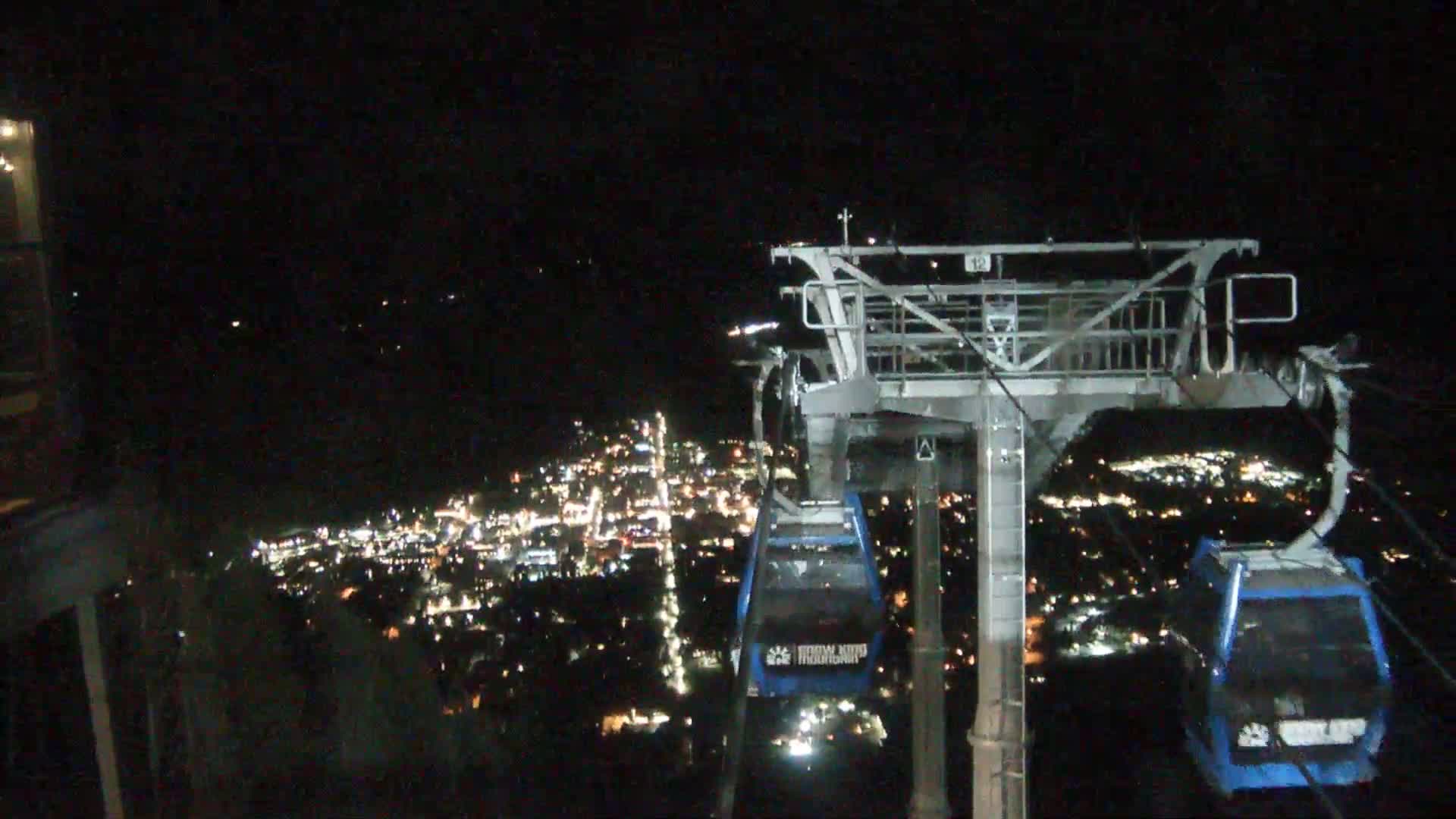 At night, a cable car ascends, passing over a brightly lit city below.