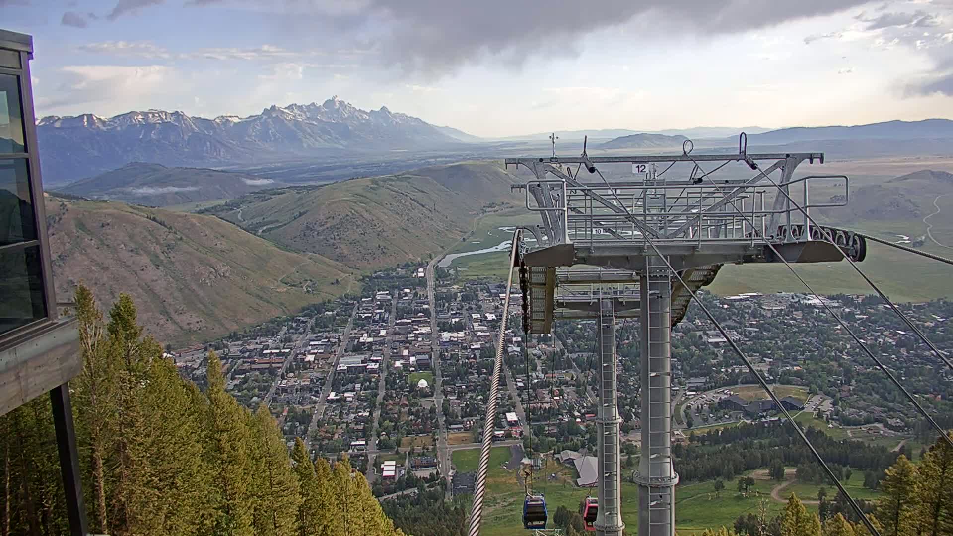 A chairlift towers over a town nestled in a valley, with snow-capped mountains visible in the distance under a partly cloudy sky.