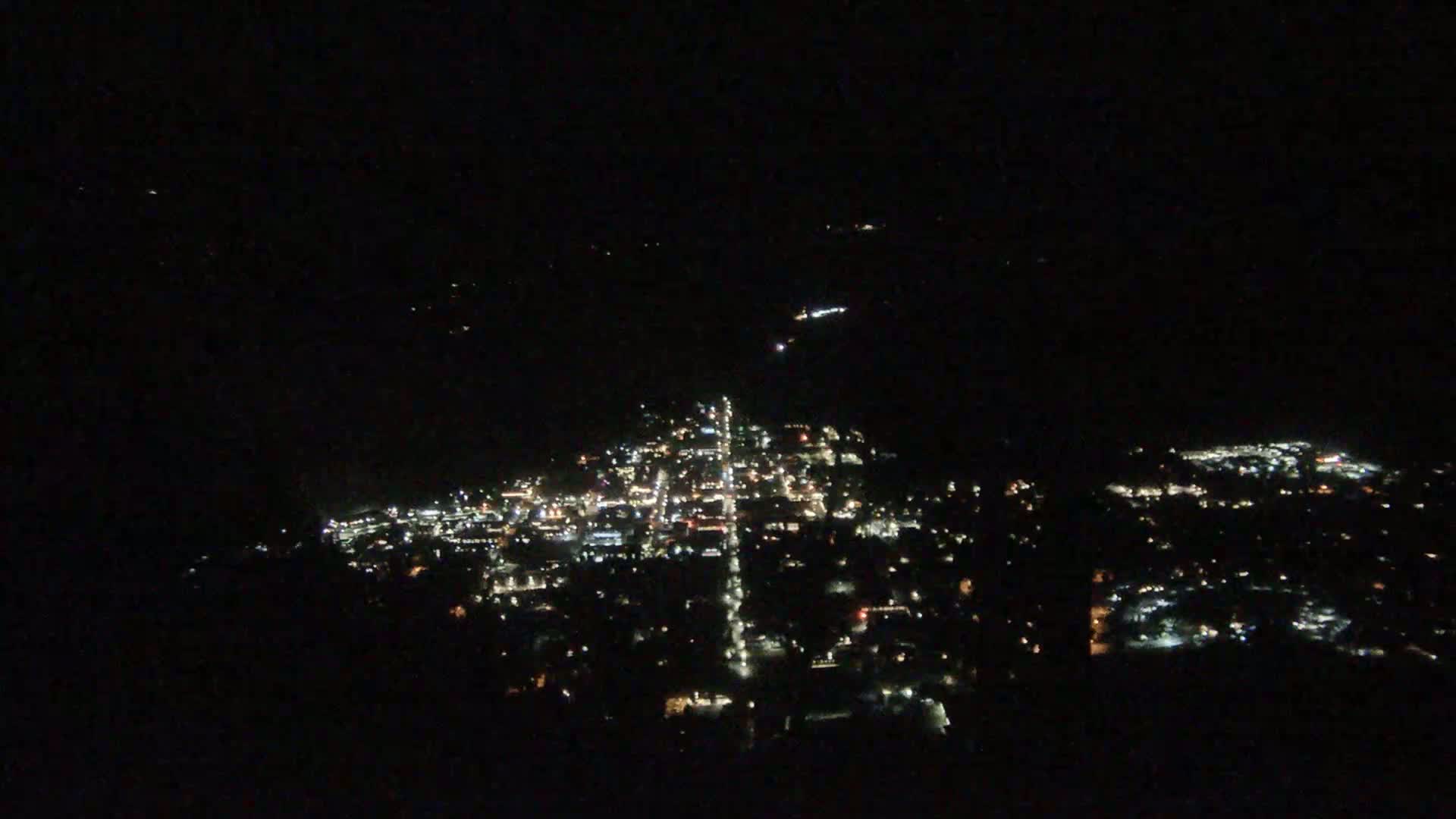 A nighttime, aerial view shows a town's illuminated streets and buildings spread across a dark landscape.