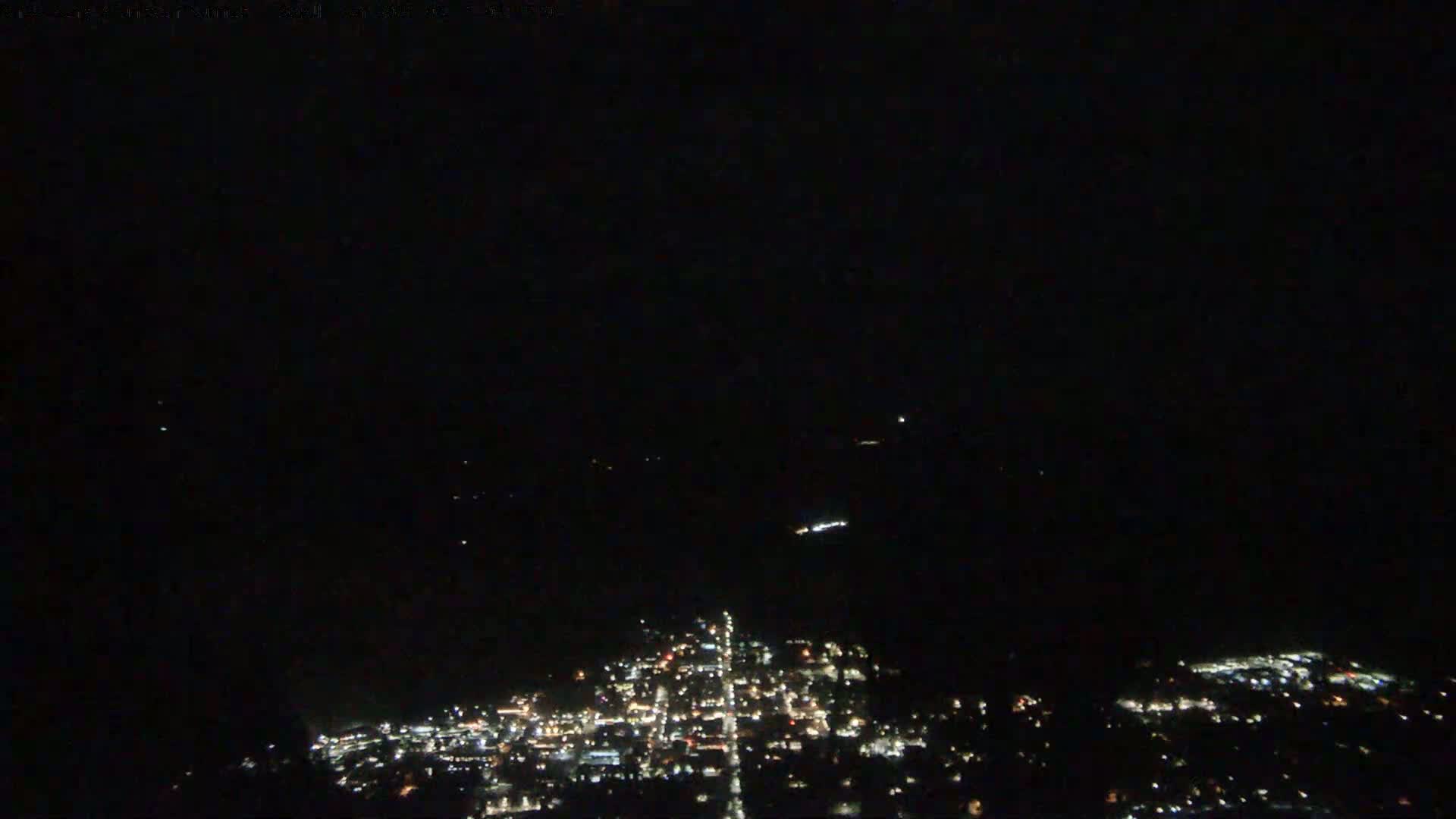 A nighttime, aerial view shows a town's illuminated streets and buildings spread across a dark landscape.