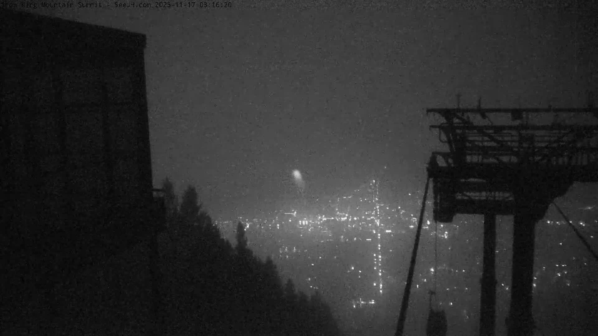 From a mountain summit at night, a grainy scene shows a city's lights faintly visible through a hazy, overcast atmosphere with a prominent plume of light rising above, framed by a dark building and a tram tower in the foreground.