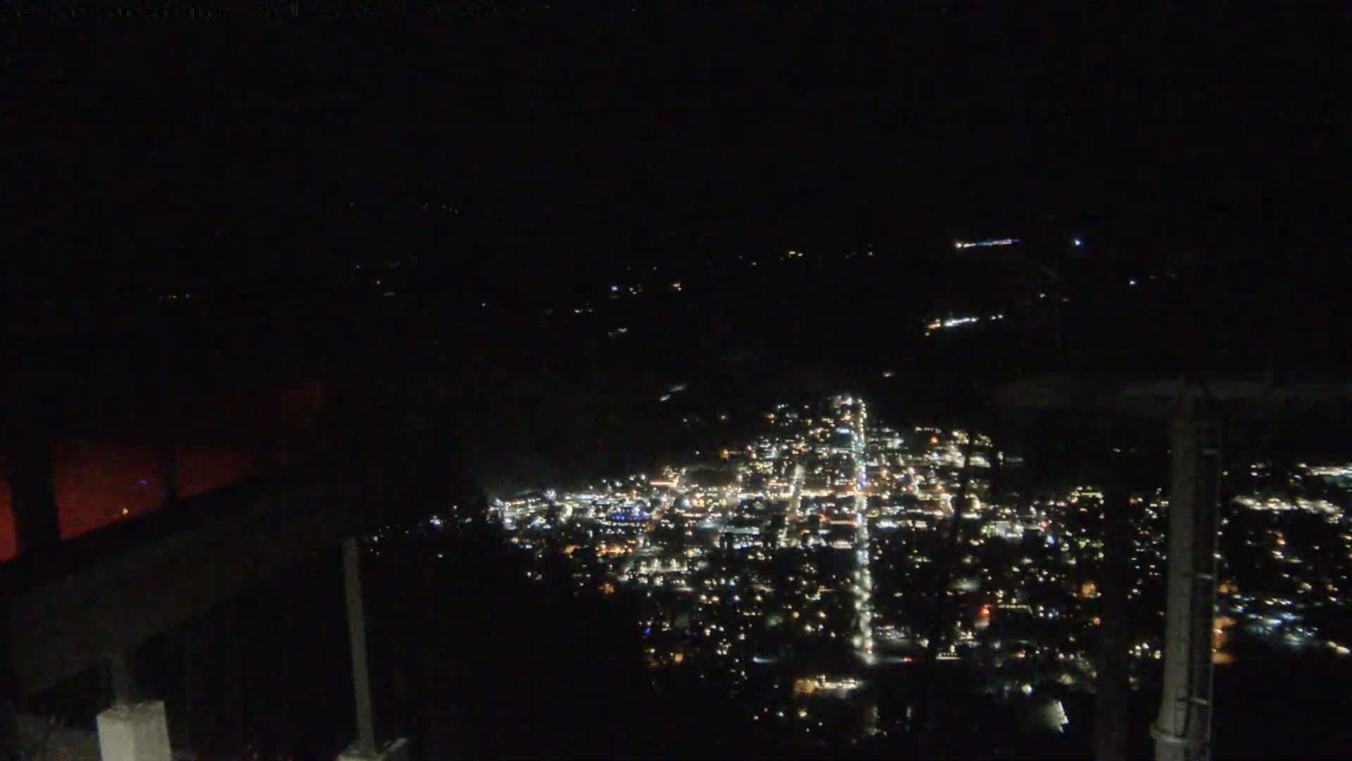 From a mountain summit at night, a grainy scene shows a city's lights faintly visible through a hazy, overcast atmosphere with a prominent plume of light rising above, framed by a dark building and a tram tower in the foreground.