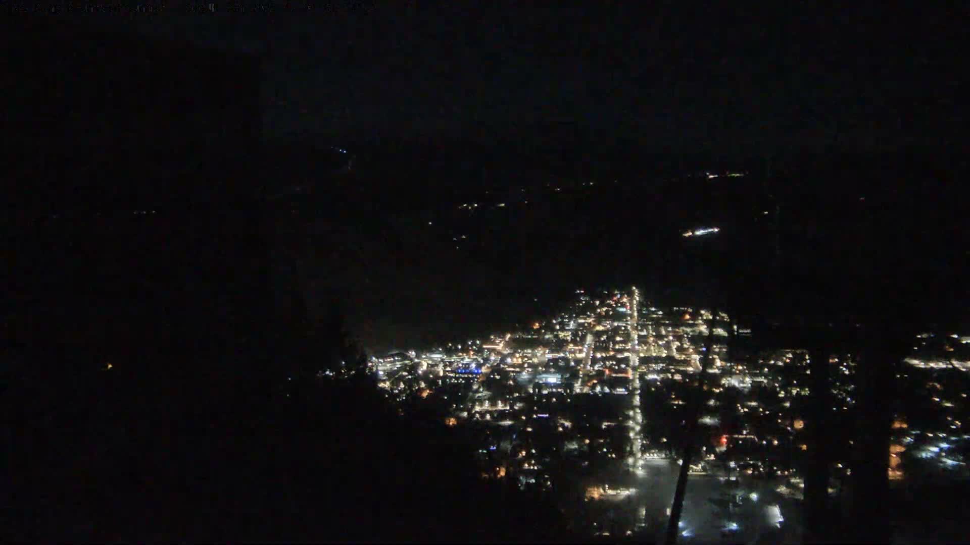 A clear night view from above shows a brightly lit city with visible streets and buildings, nestled in a dark, mountainous landscape.