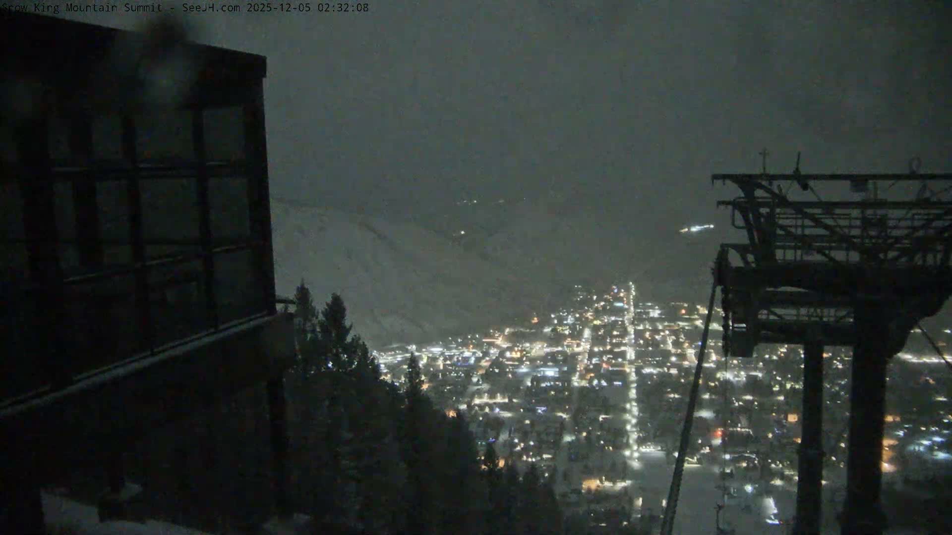 A panoramic night scene from a snowy mountain reveals a brightly lit town in the valley below, with dark foreground structures including a building and a ski lift tower, all under an overcast sky with visible snowfall.