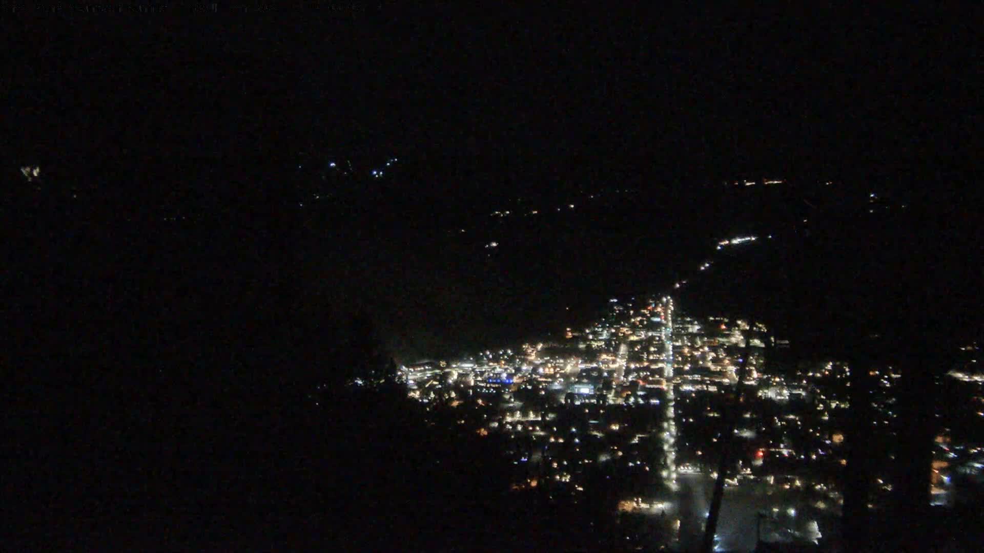 A panoramic night scene from a snowy mountain reveals a brightly lit town in the valley below, with dark foreground structures including a building and a ski lift tower, all under an overcast sky with visible snowfall.