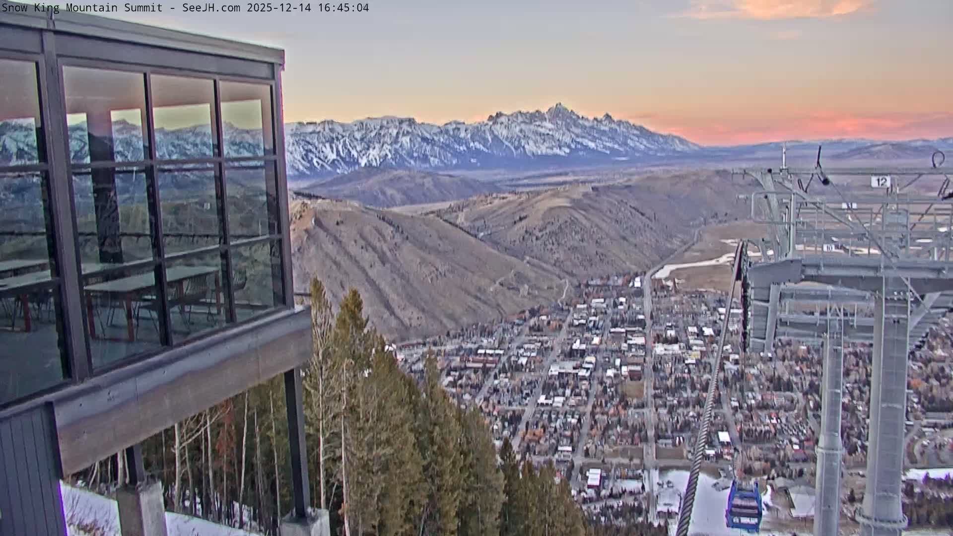 A panoramic night scene from a snowy mountain reveals a brightly lit town in the valley below, with dark foreground structures including a building and a ski lift tower, all under an overcast sky with visible snowfall.