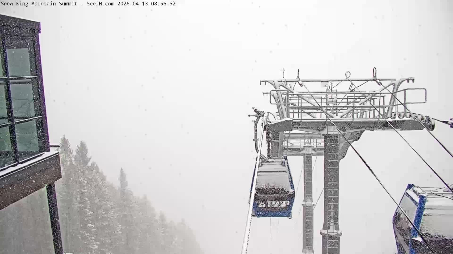 A panoramic night scene from a snowy mountain reveals a brightly lit town in the valley below, with dark foreground structures including a building and a ski lift tower, all under an overcast sky with visible snowfall.