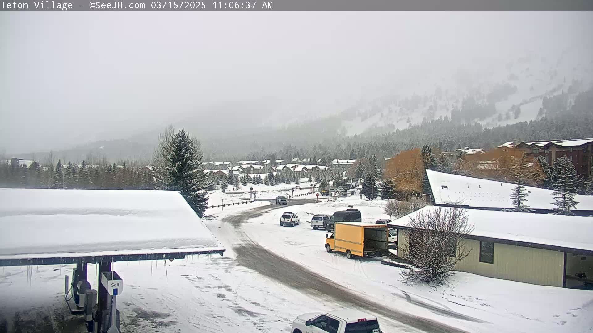 A snow-covered village nestled in a mountain valley during a light snowfall, with several vehicles parked along a road.