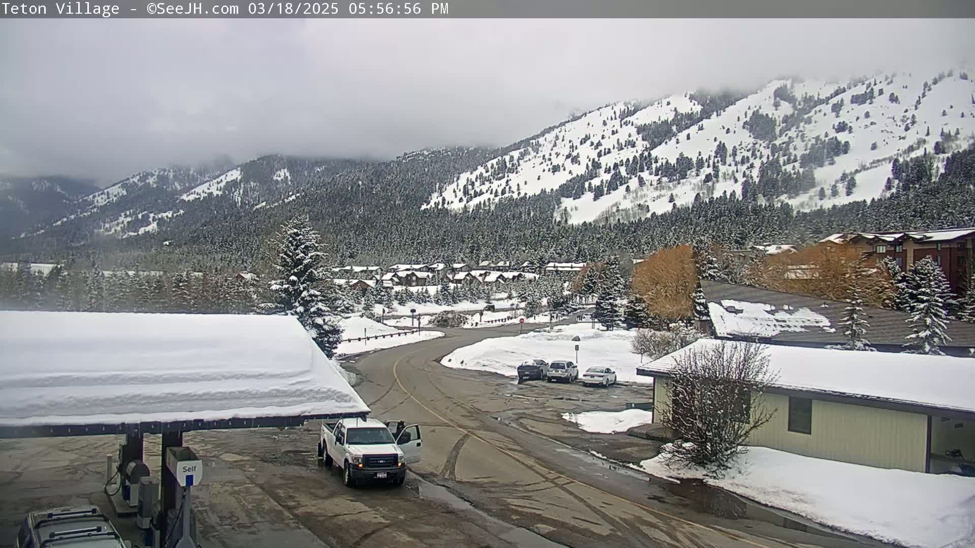 A snow-covered village nestled in a valley beneath a snow-dusted mountain range on an overcast day.