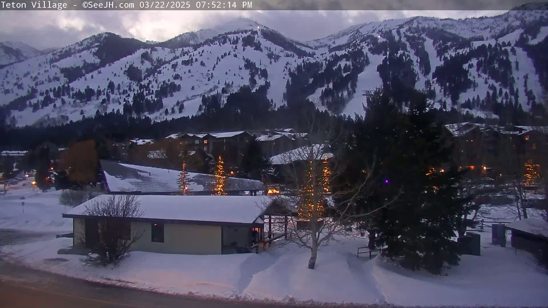 A snow-covered village at dusk is nestled at the base of a snow-dusted mountain range.