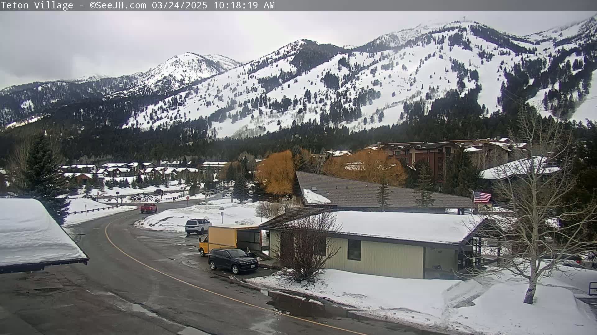 A snow-covered mountainside overlooks a residential area with several vehicles parked near a small building on a partly cloudy day.