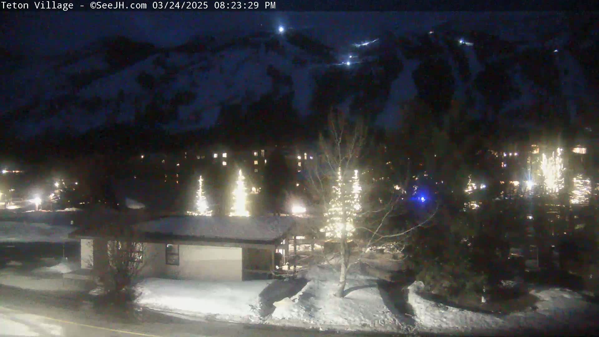 A nighttime, snow-covered scene shows a village nestled below a dark mountain, illuminated by lights from buildings and trees decorated with lights.