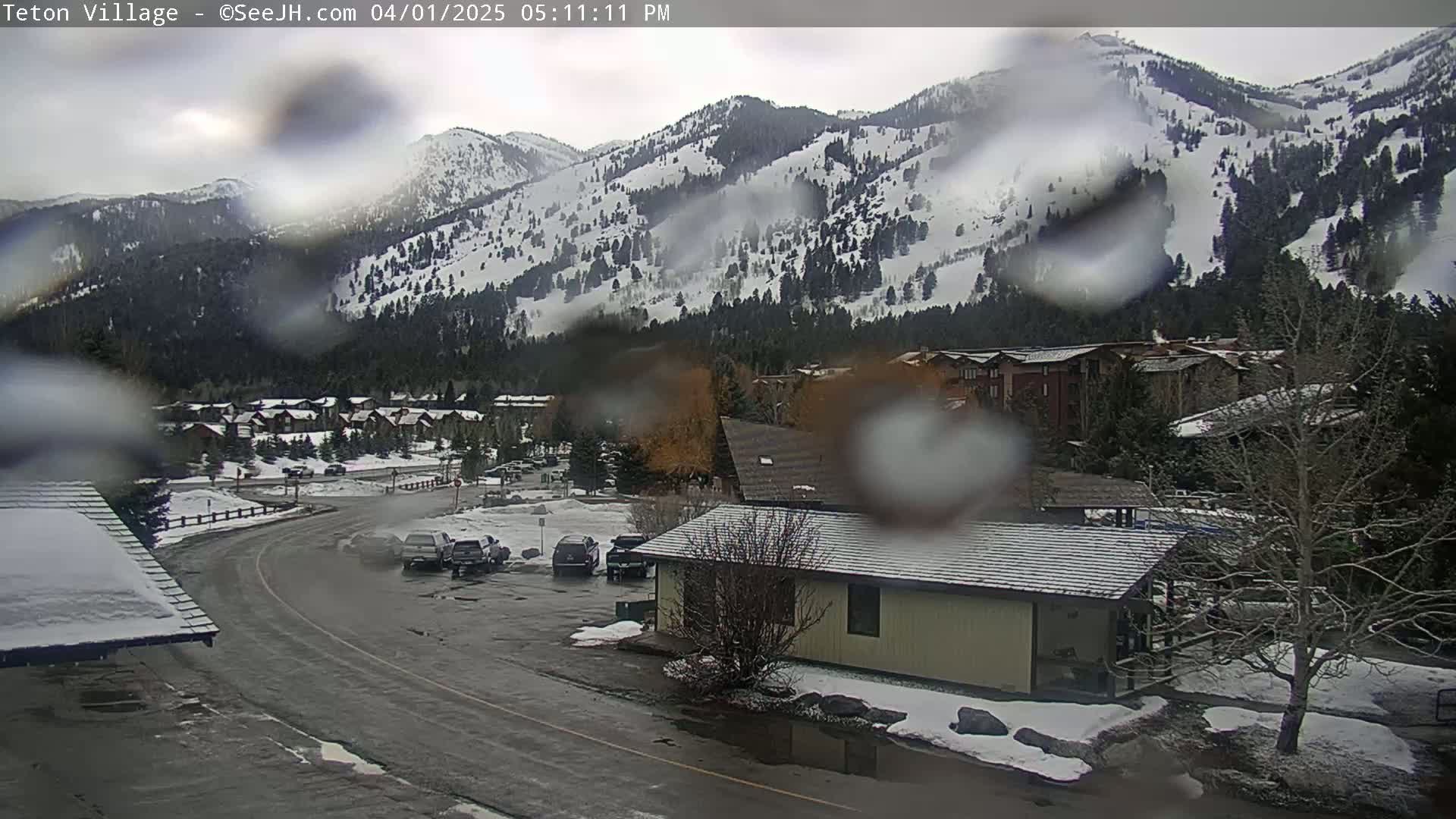 A rain-covered view shows a snow-dusted mountainside overlooking a village with parked cars and a small building in the foreground.