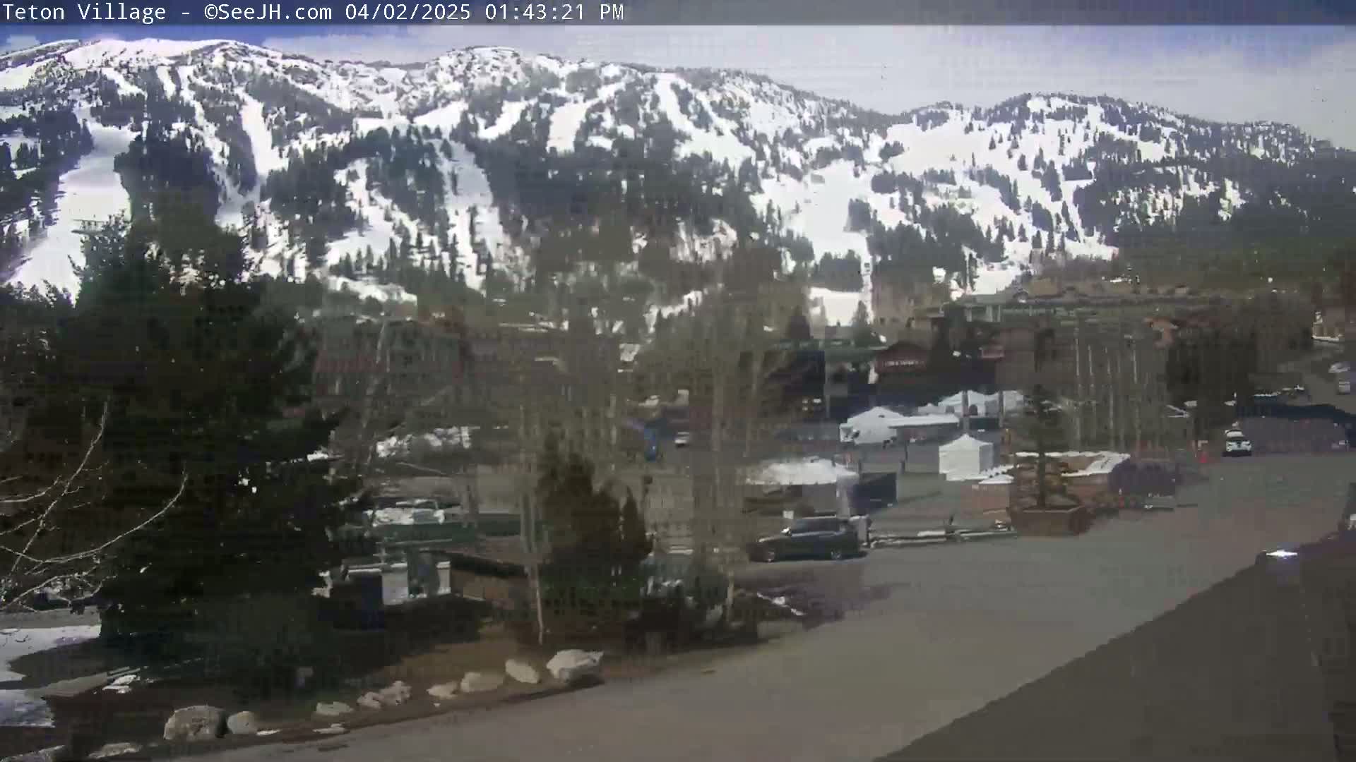 A partly cloudy day shows a snow-covered mountainside overlooking a village with buildings, cars, and trees.
