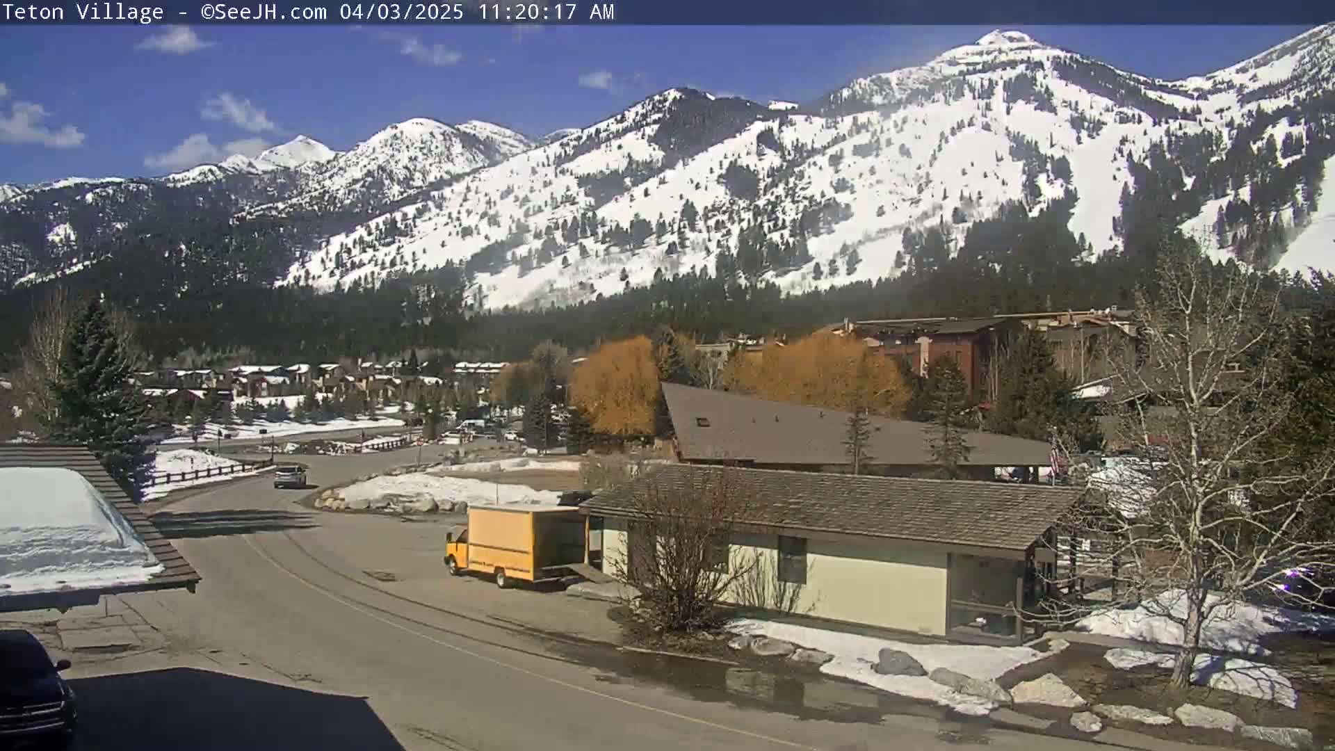 A sunny day reveals a snow-capped mountain range overlooking a small town with buildings and vehicles, partially snow-covered ground, and some bare trees.