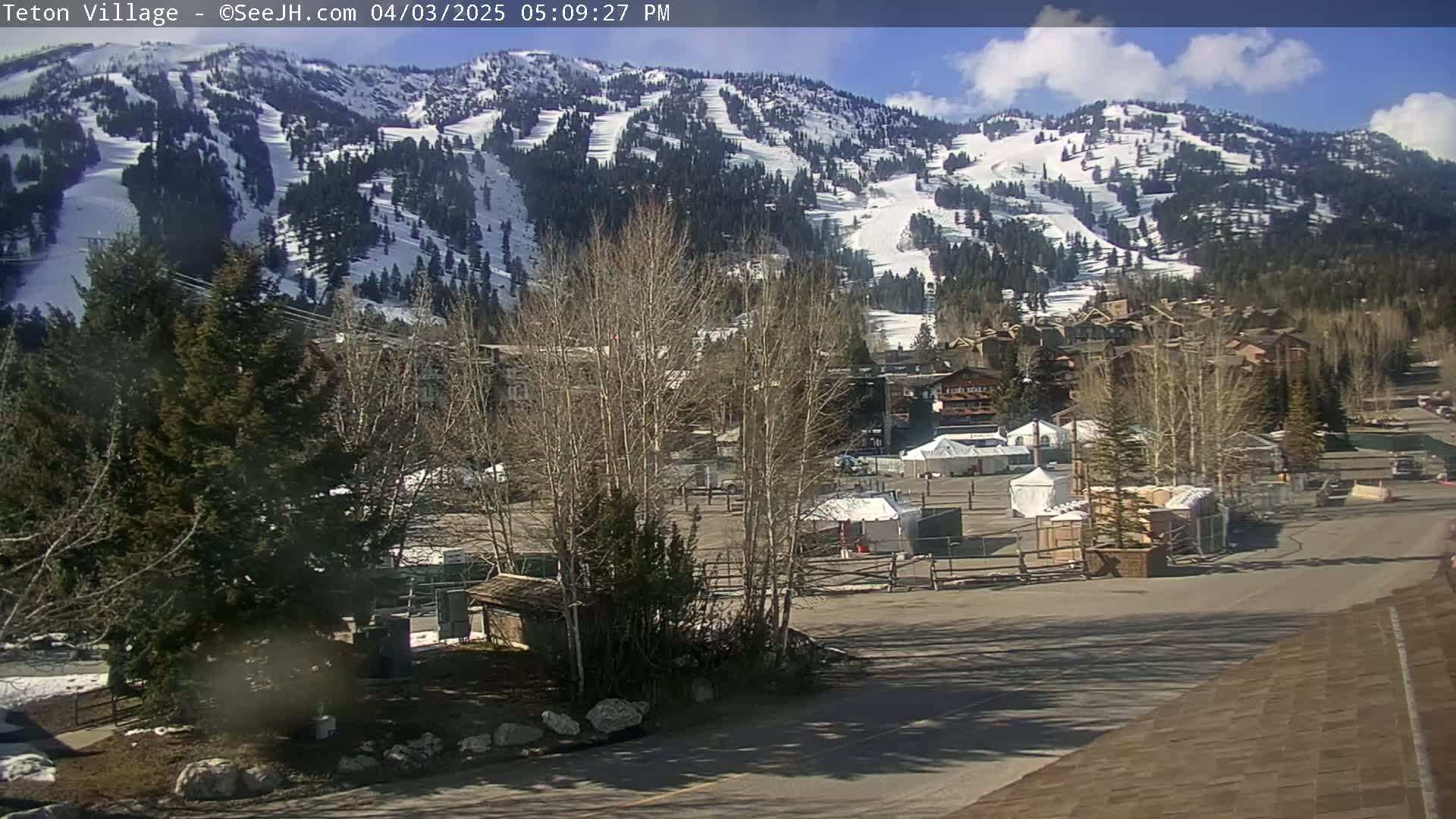 A snow-covered mountainside with ski runs is visible behind a village of buildings and a mostly empty parking lot on a sunny day.