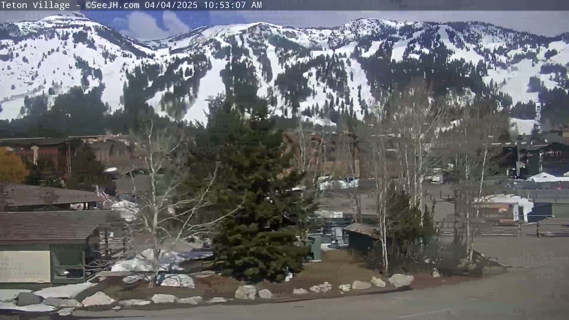 A snow-covered mountain range is visible in the background, under a partly sunny sky, behind a village of buildings and trees.