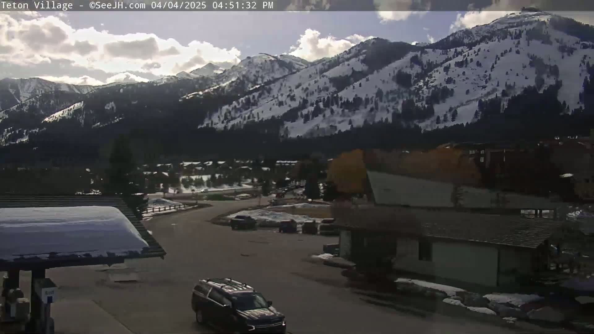 A partly cloudy day reveals snow-covered mountains in the background, overlooking a parking lot with several vehicles and buildings in the foreground.