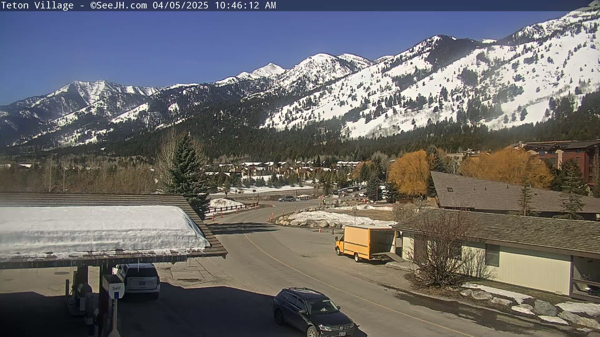 A sunny day reveals a snow-covered mountain range behind a village with buildings and vehicles, partially snow-covered roads, and a gas station canopy with some remaining snow.
