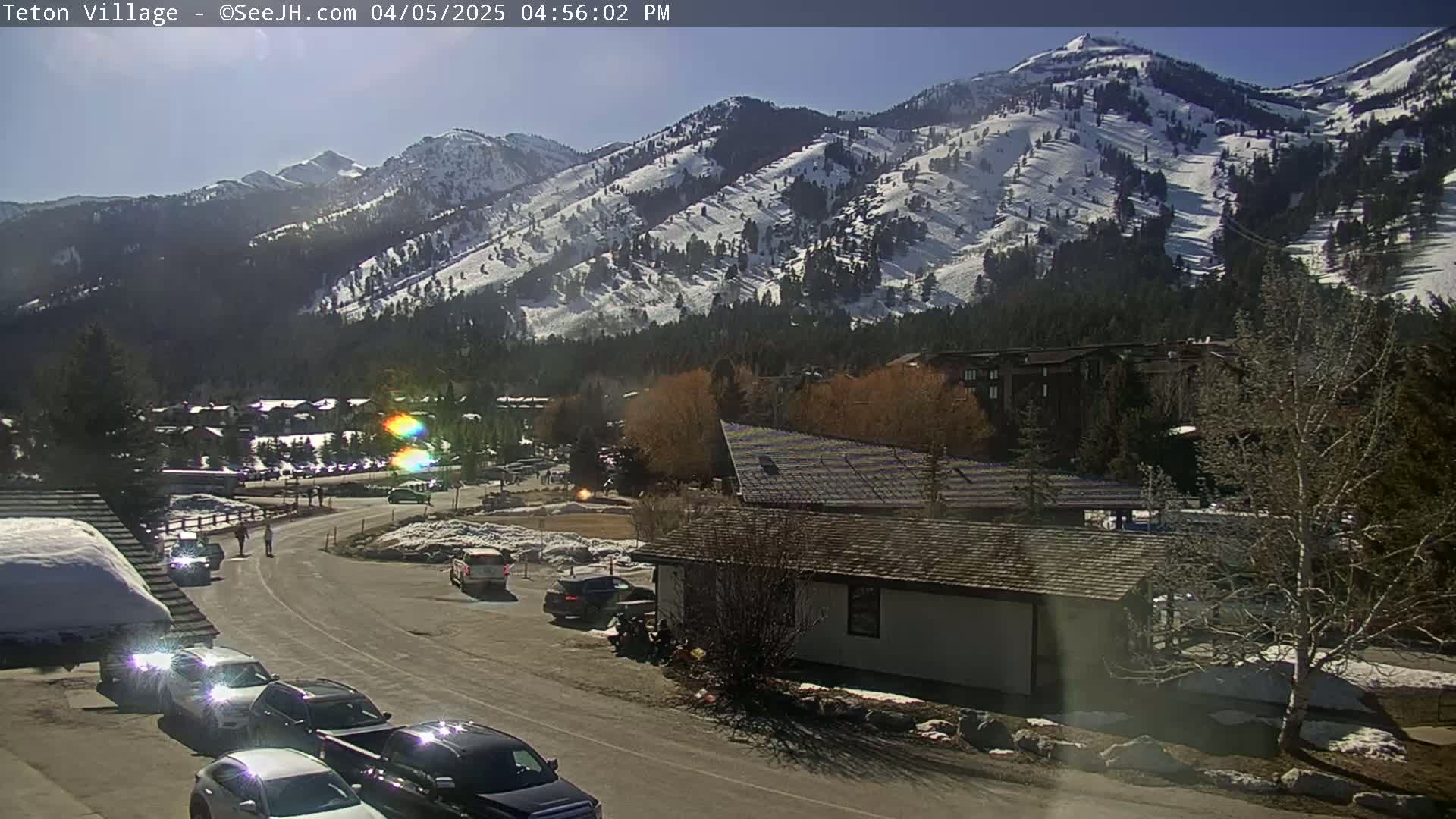 A sunny day reveals a snow-covered mountain range overlooking a parking lot and road with cars, a small building, and some pedestrians in a village setting.