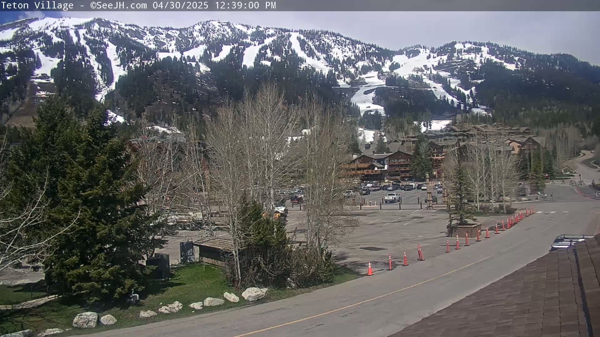 A partially snow-covered mountain is visible behind a parking lot and buildings, under a mostly sunny sky.