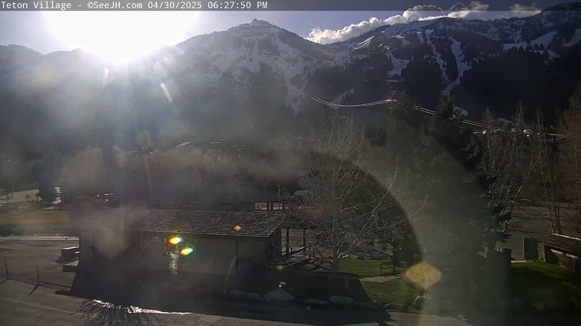 A sunny, hazy afternoon view shows a small building and bare trees in the foreground with snow-capped mountains in the background.