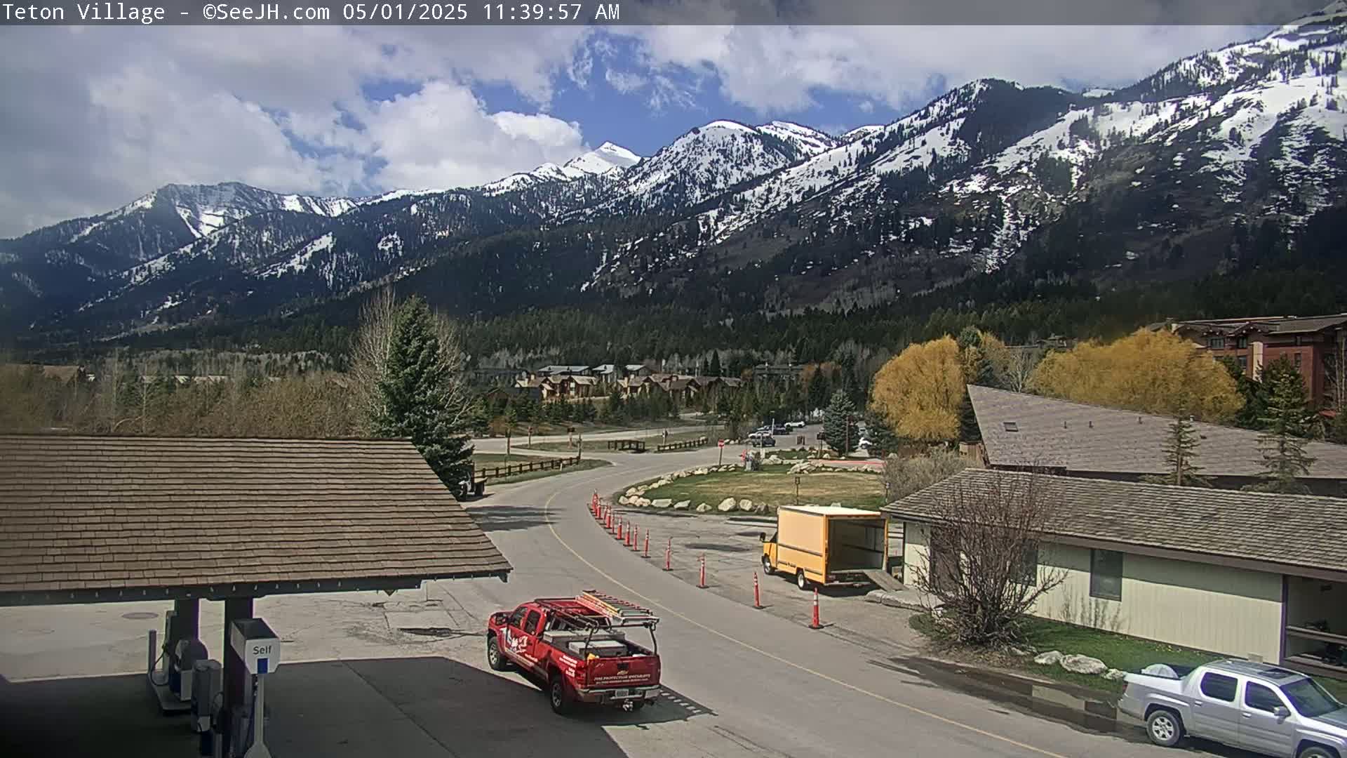 A partly cloudy day reveals snow-capped mountains overlooking a village with roads, buildings, and several vehicles, including a red truck and a yellow van.