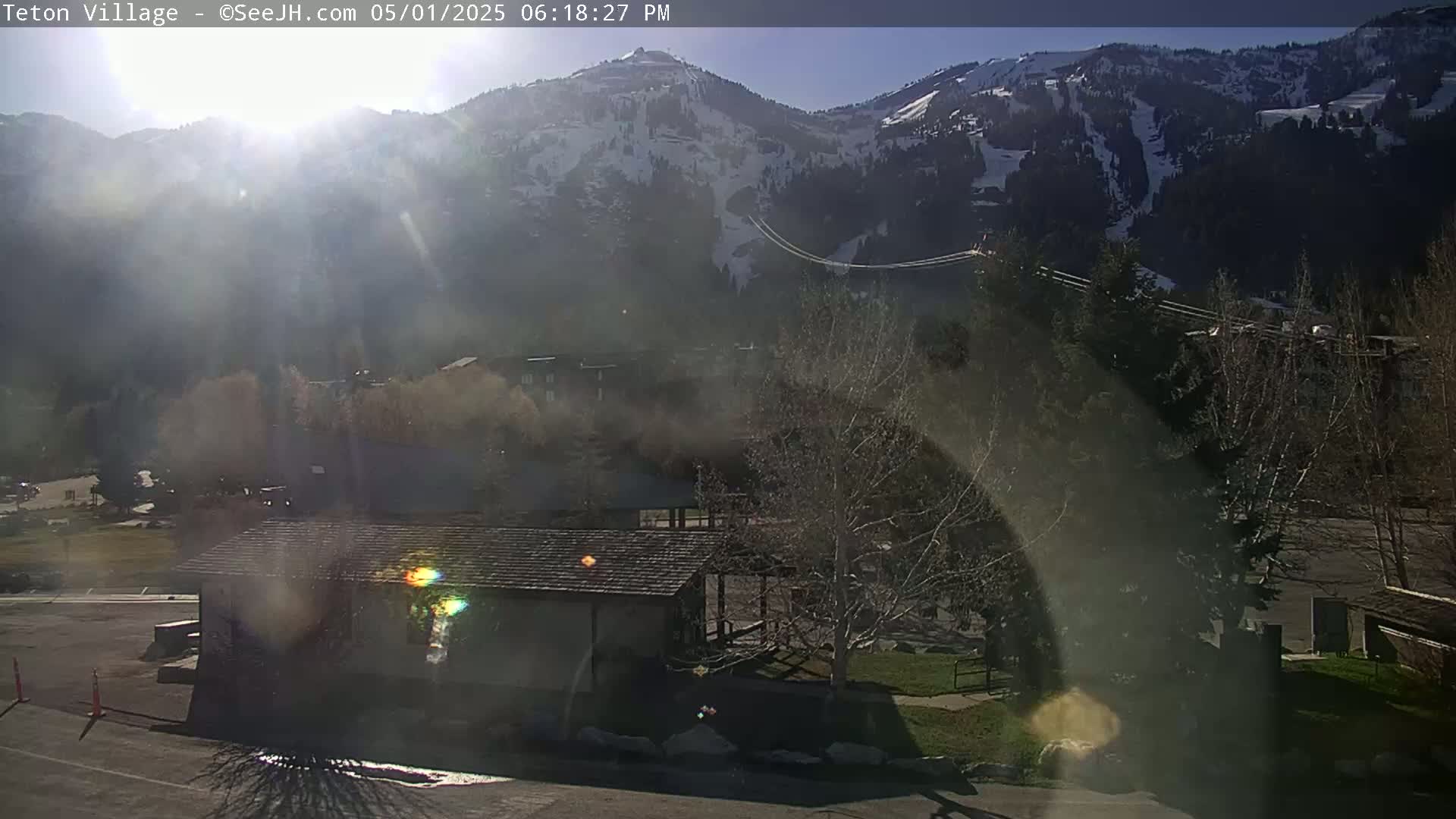 A hazy, sunny day reveals a snow-capped mountain range behind a small building and leafless trees in a village setting.