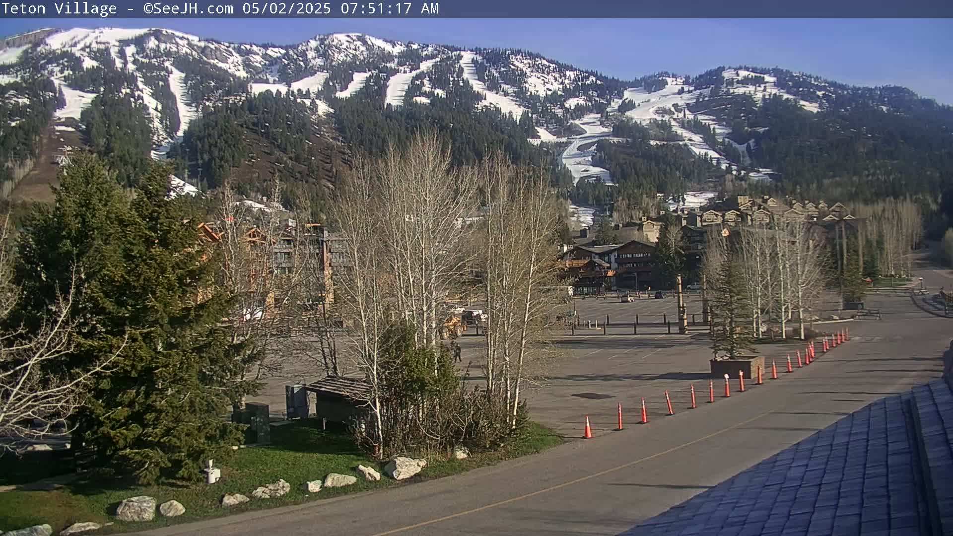 A mostly empty parking lot sits in front of buildings and trees, with a snow-dusted mountain in the background under a sunny sky.