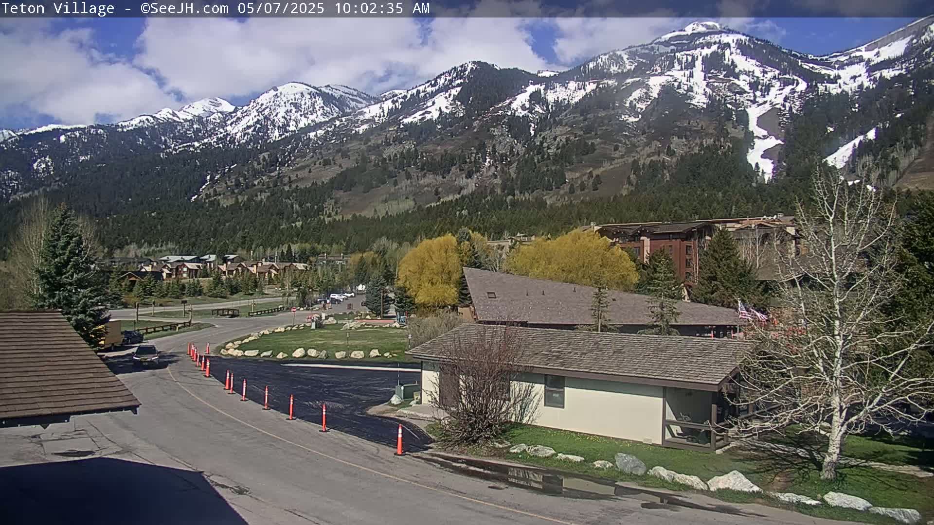 A partly sunny day reveals snow-capped mountains in the background, overlooking a village with buildings, roads, and some yellow-leafed trees, with fresh asphalt visible in the foreground.