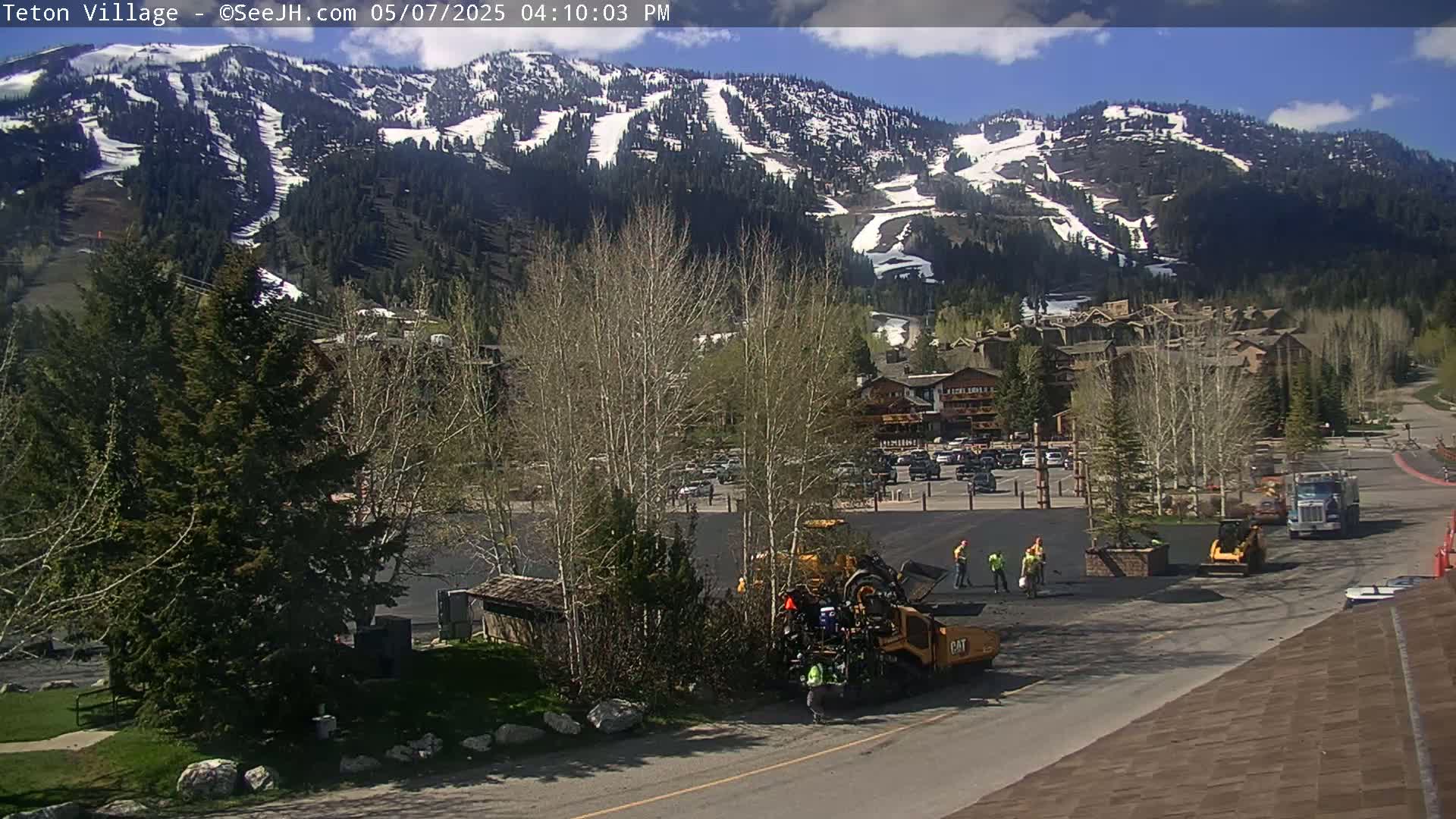 A sunny day shows road construction crews paving a parking lot in a village nestled at the base of a snow-capped mountain range.