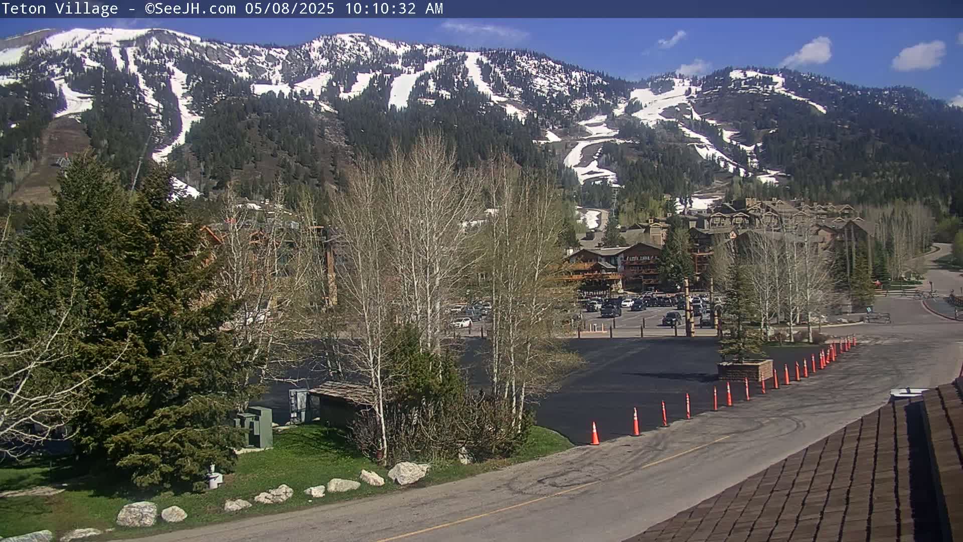 A mountainside with snow patches is visible behind a parking lot and buildings under a mostly sunny sky.