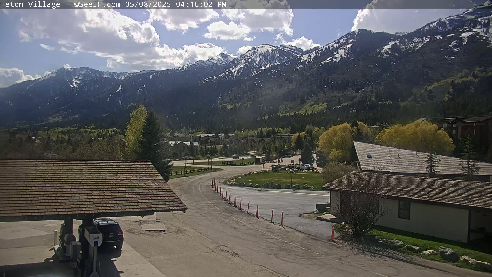 A partly cloudy day reveals a mountain range with some snow, overlooking a village with buildings and a paved road.