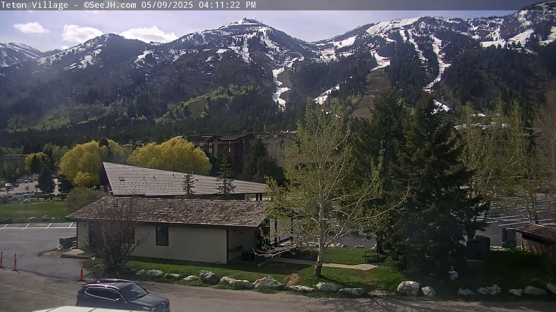 A partly sunny day reveals a snow-capped mountain range overlooking a residential area with a small building and a car parked nearby.