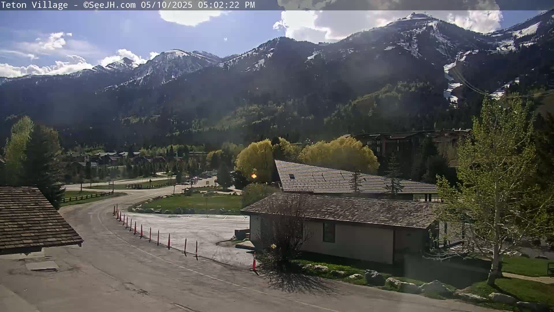 A partly sunny day reveals a mountain village nestled at the base of snow-capped mountains, with a road curving towards a small building.