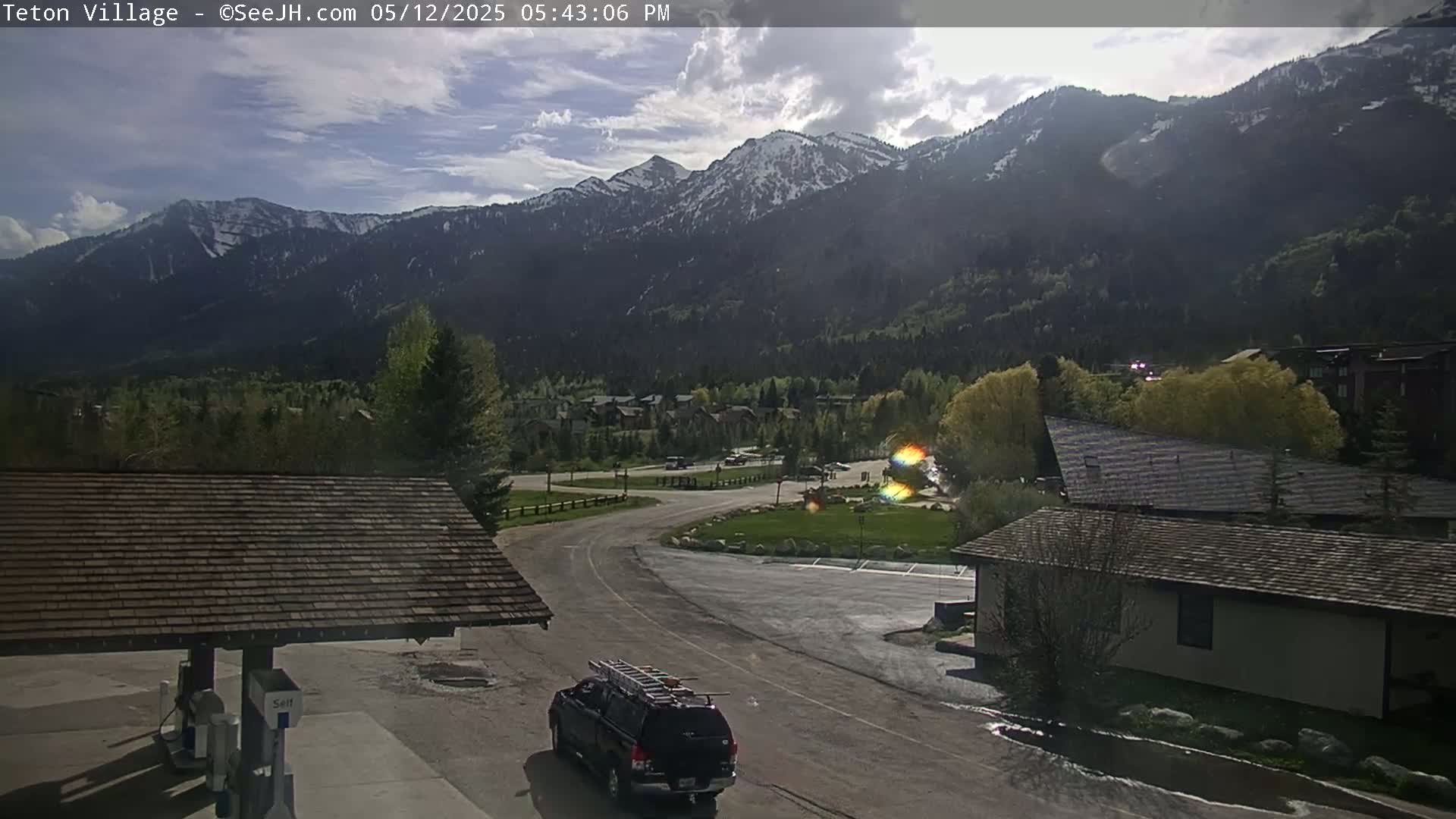 A partly cloudy afternoon scene shows a mountain range with some snow, a road curving through a village with several buildings and a dark-colored SUV, all under a mostly sunny sky.
