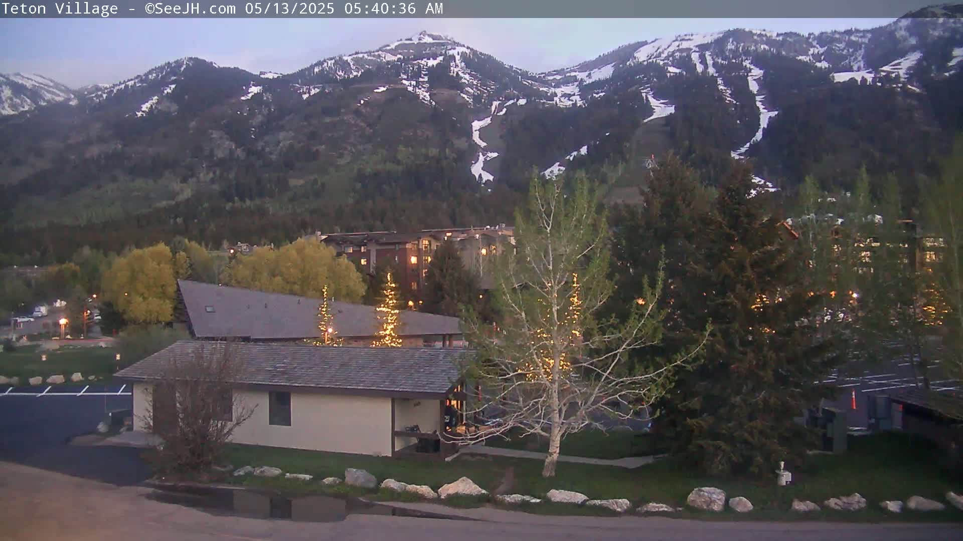 A low-light view of a small building and parking lot nestled amongst trees, with snow-capped mountains in the background under a twilight sky.
