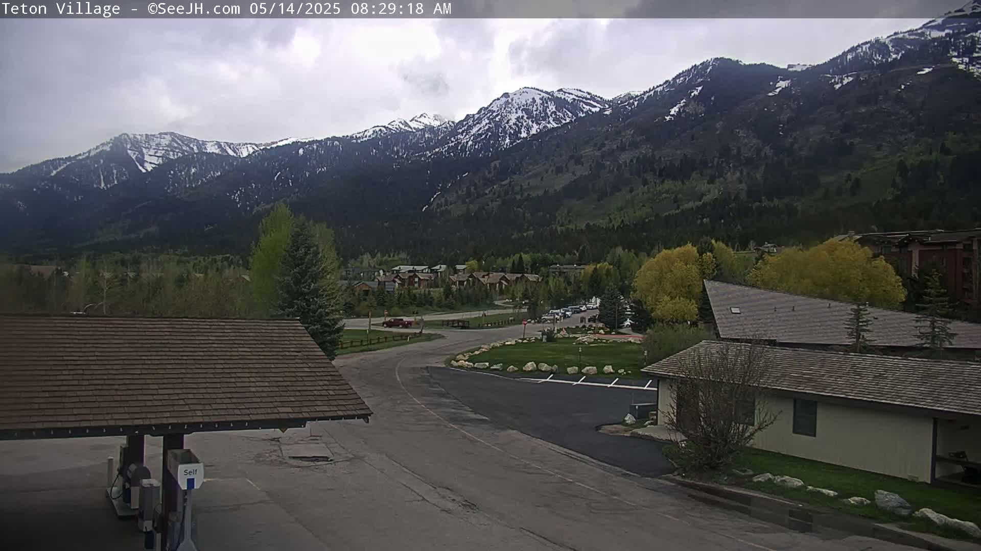 A view of a mountain range with snow-capped peaks, partially overcast skies, and a small village nestled in the valley below.