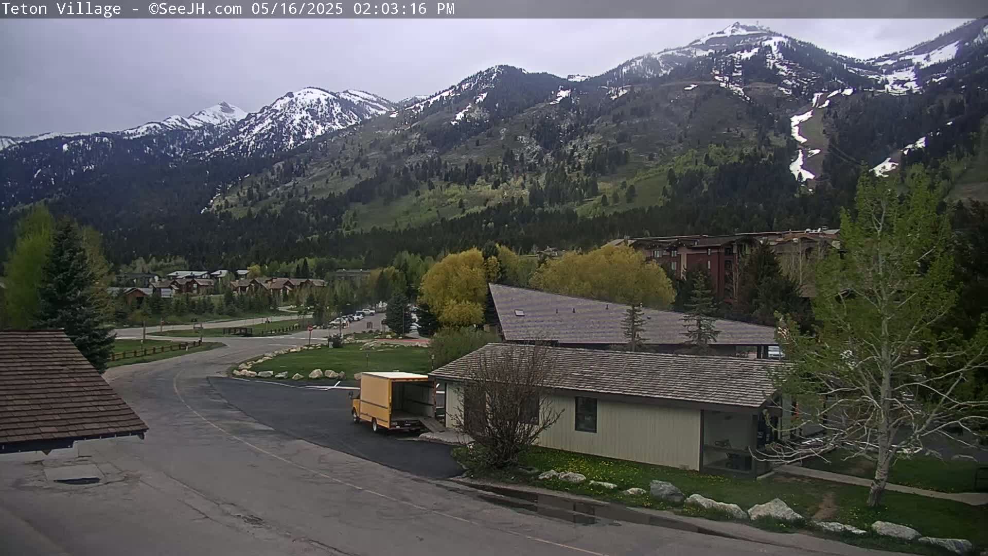 A partially cloudy day reveals a mountainous landscape with snow-dusted peaks, a small town nestled in the valley below, and a delivery truck parked near a low building.