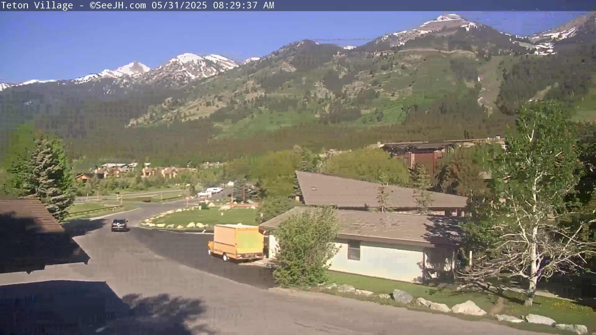 A sunny day shows snow-capped mountains in the background, with a small town and a few vehicles visible in the foreground.